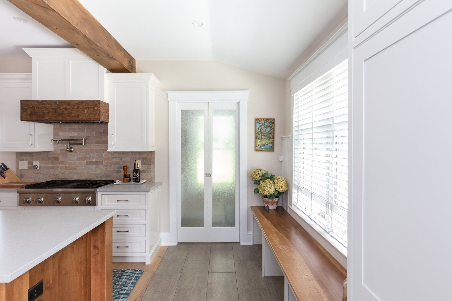 Bright modern kitchen with white cabinets, wood accents, and a window seat by large windows.
