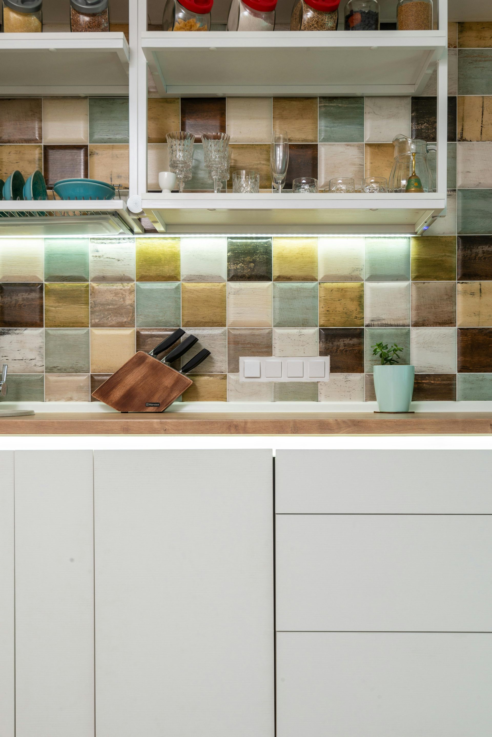 Kitchen with multicolored square tile backsplash, shelves, white cabinets, wooden countertop.