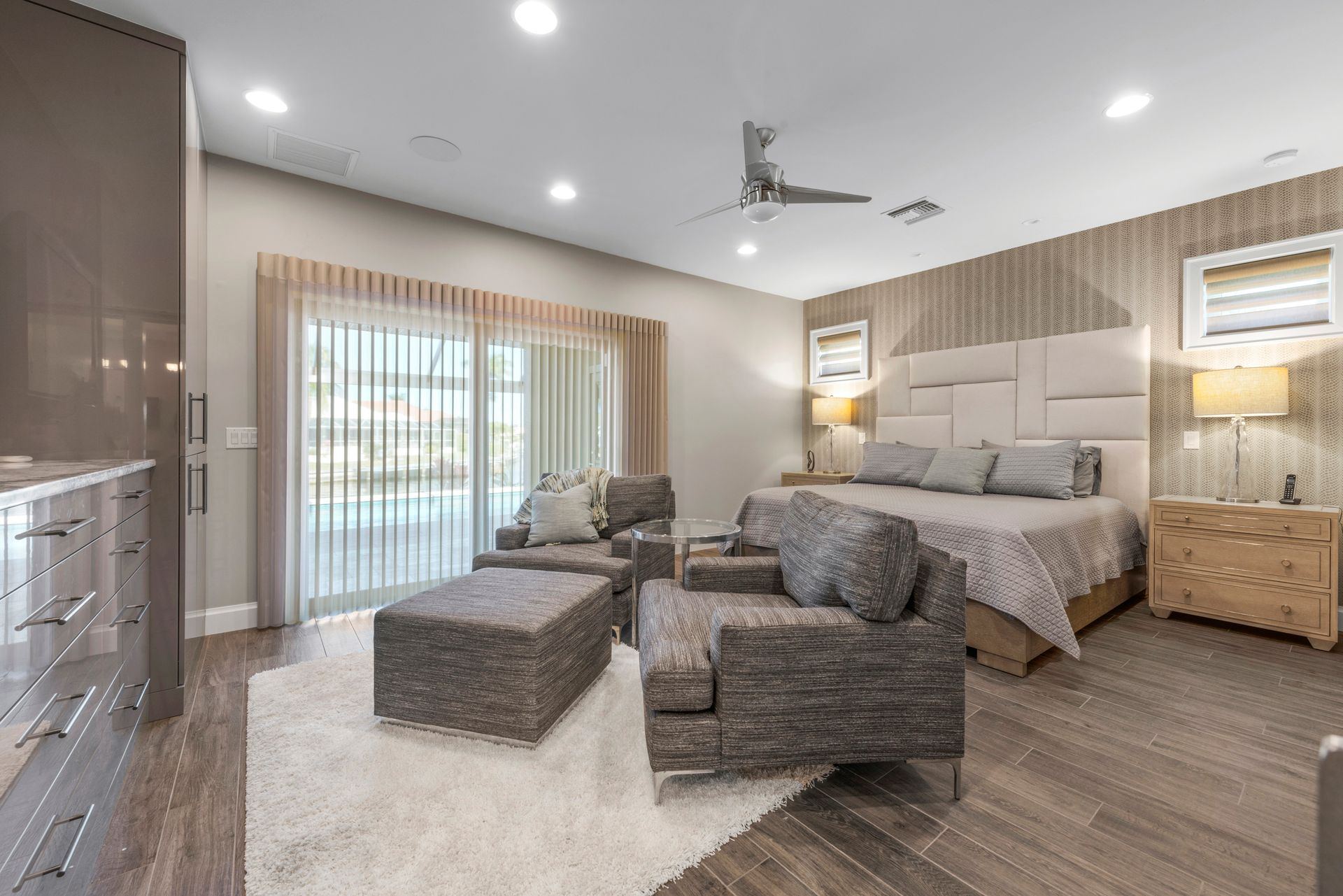 A large bedroom with a king size bed , chairs , ottomans and a ceiling fan.