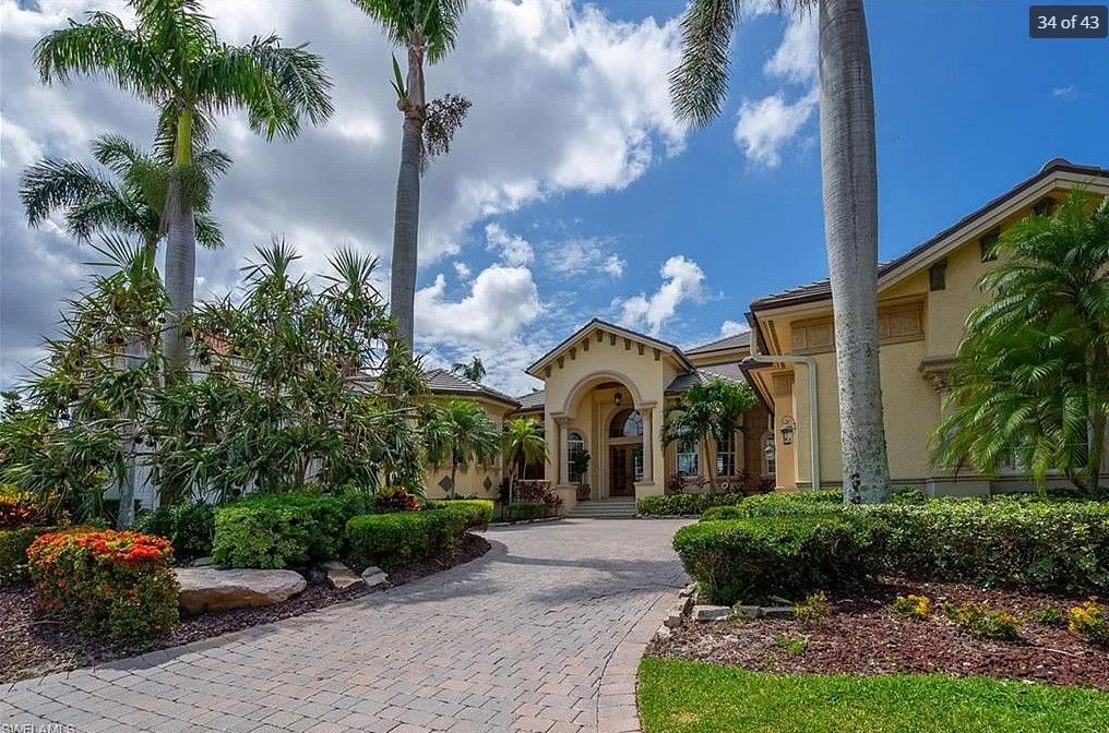 A stately, yellow stucco house with a brick driveway, framed by palm trees and lush greenery, under a blue sky.