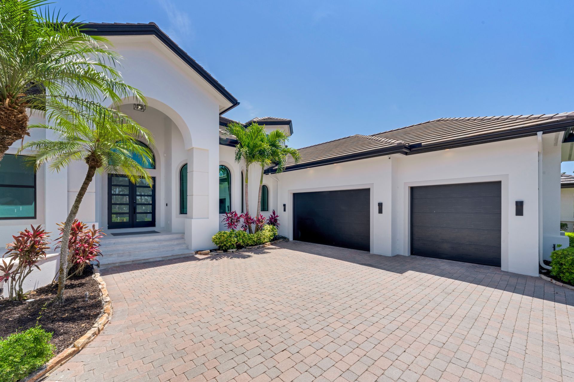 White stucco house with black garage doors and a brick driveway under a sunny sky.