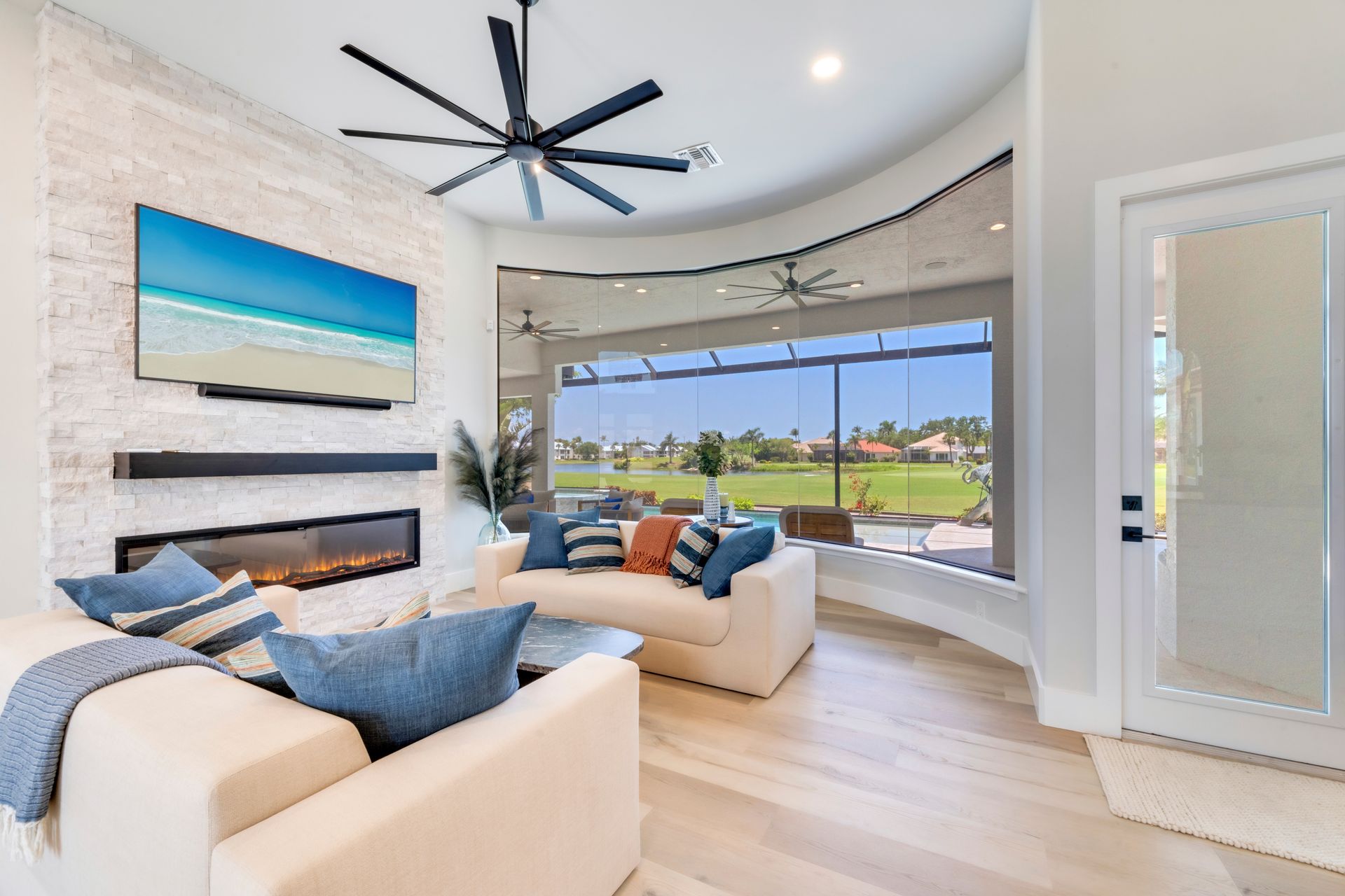 Living room with white sofas, large windows, fireplace, and TV displaying a beach scene.