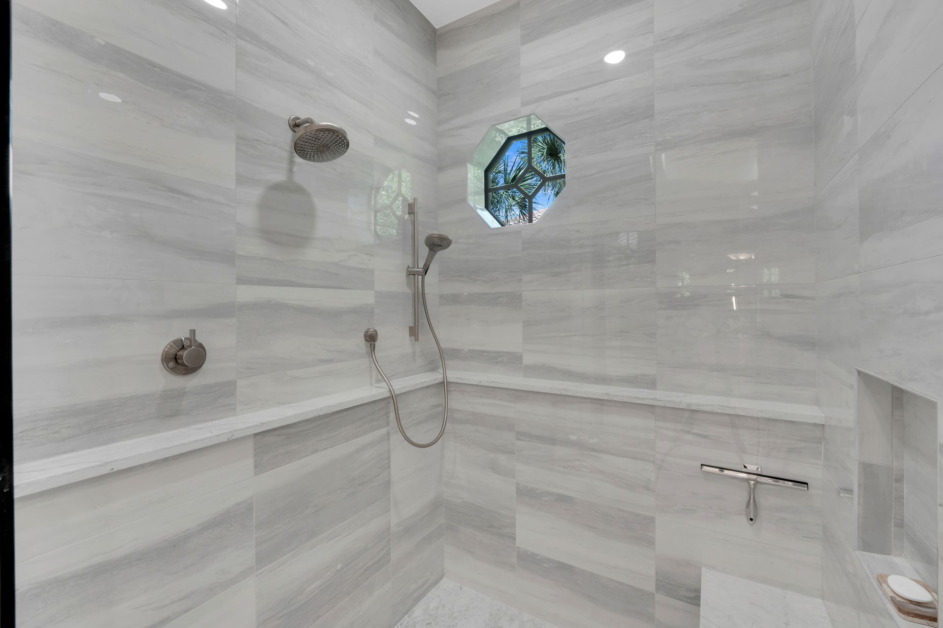 Gray tiled shower with a round window and two shower heads.