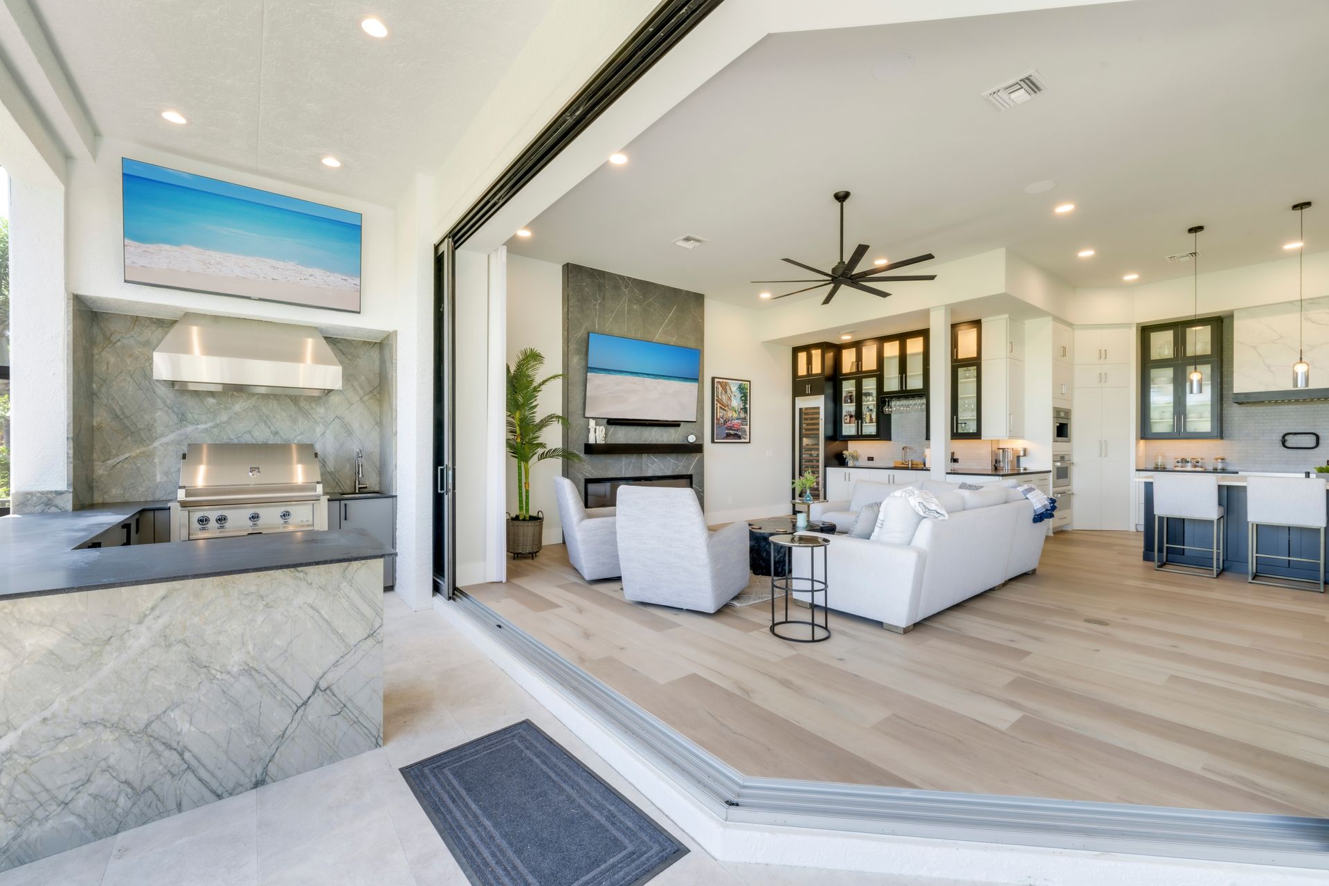 Indoor/outdoor living space with kitchen and living room open.  Light wood floors, modern furniture, and a grill.