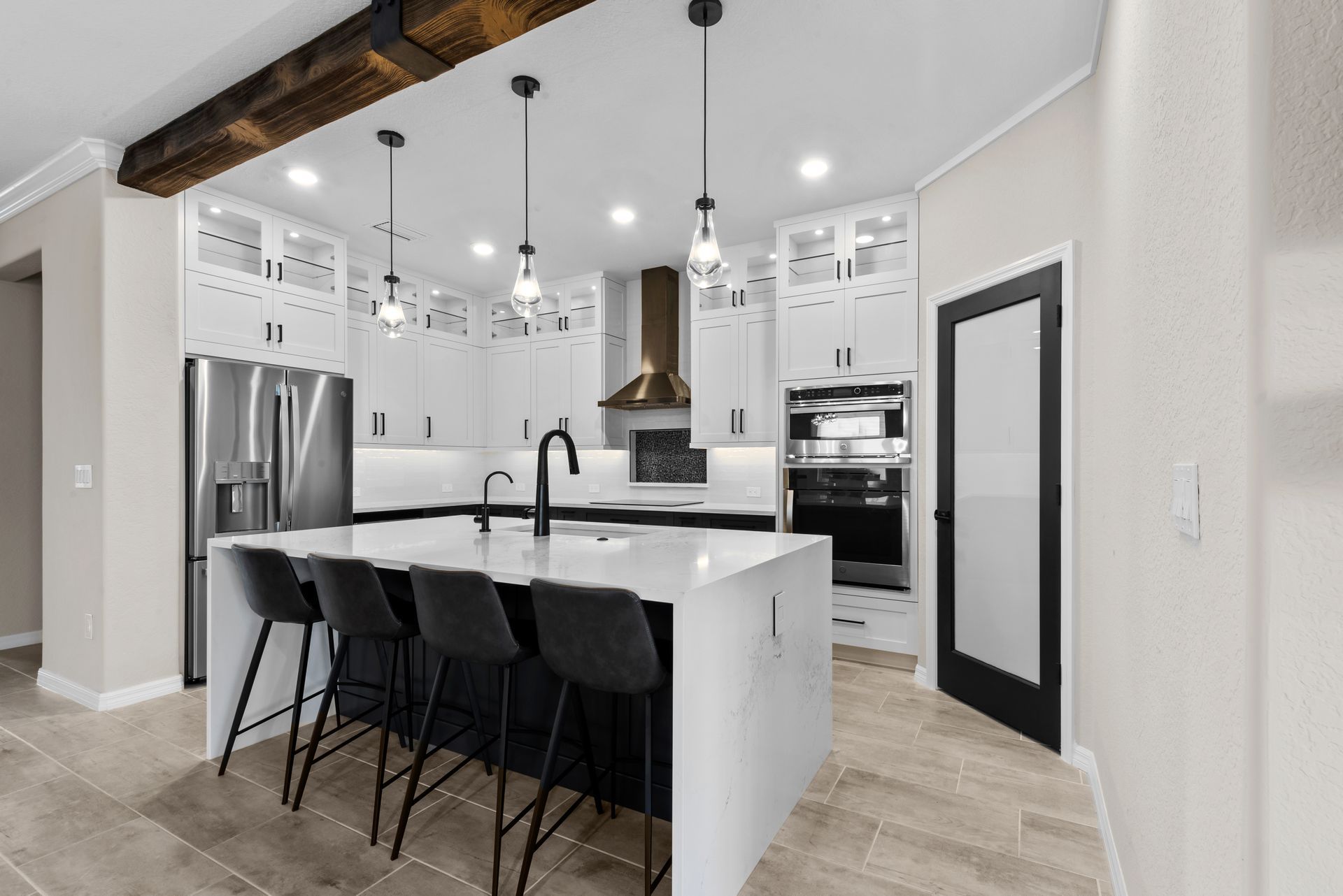 Modern white kitchen with island, black accents, stainless steel appliances, pendant lights, and exposed wood beam.