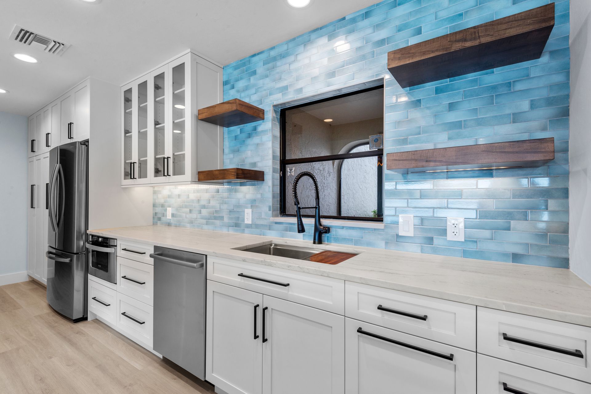 Modern kitchen with white cabinets, stainless steel appliances, blue tile backsplash, wooden shelves, and a black window frame.