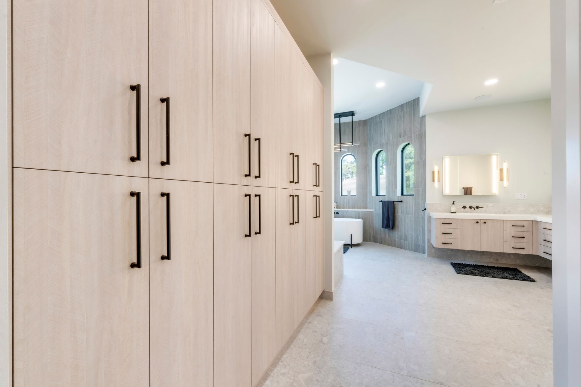 Light wood cabinetry with black handles in a bathroom, leading to a vanity and arched windows.