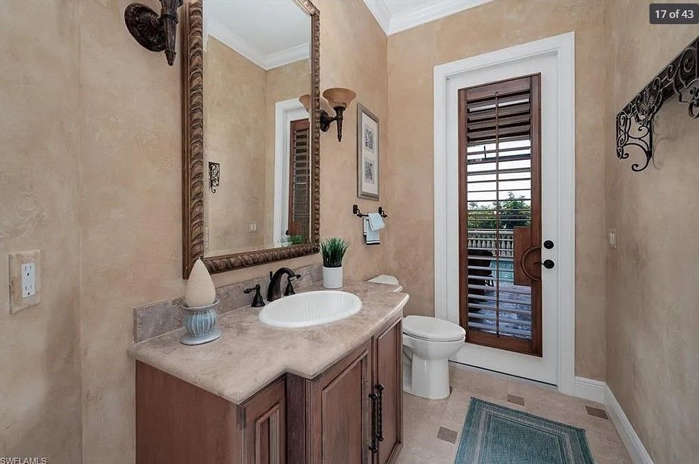 Bathroom with sink, mirror, and toilet. Beige walls, brown vanity, and a door with shutters.