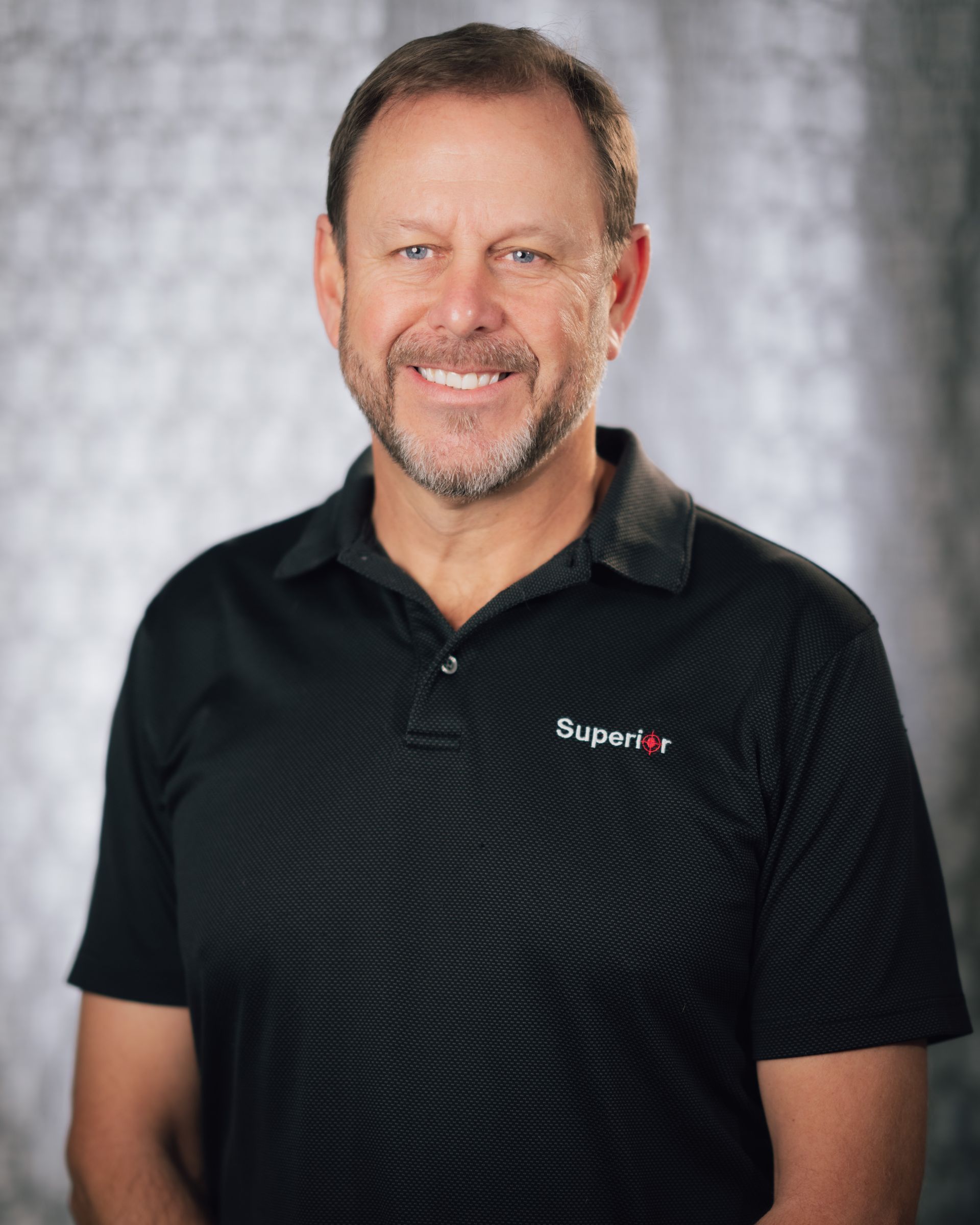 A man with a beard is wearing a black polo shirt and smiling.