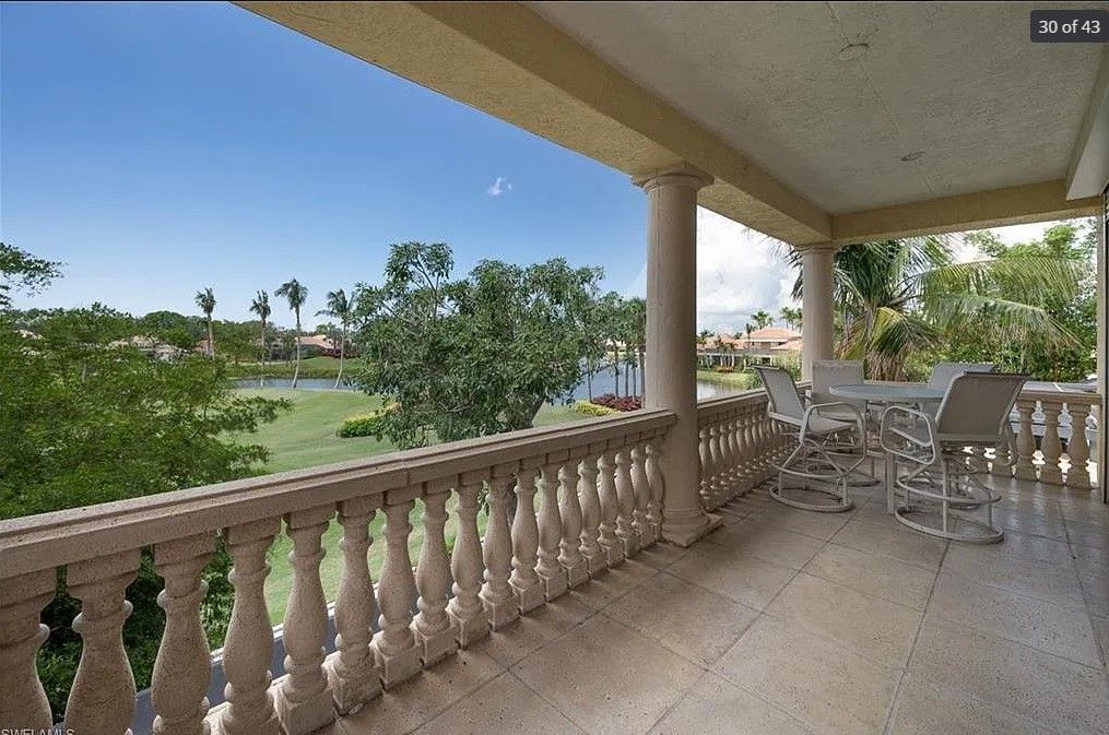 Balcony overlooking a waterway, with a table, chairs, and ornate railing. Blue sky, greenery.
