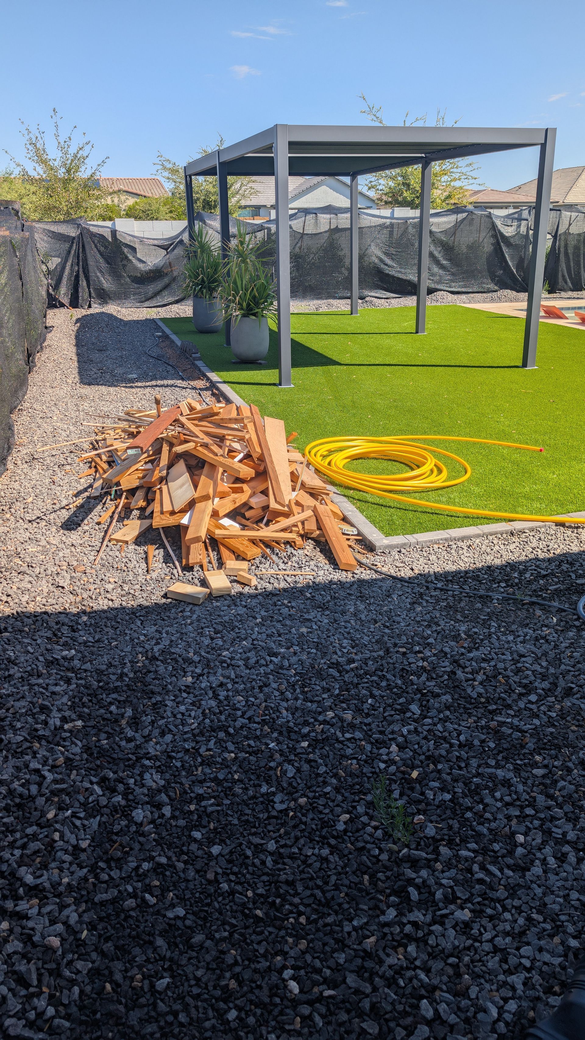 Backyard with gray pergola, artificial turf, gravel border, and wood debris.