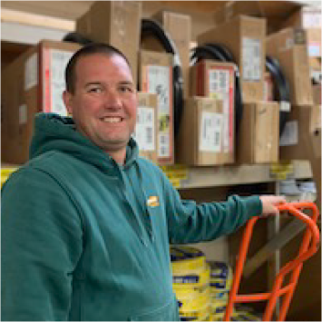 Man in green hoodie, smiling, leaning on a hand truck, boxes and shelves in the background.
