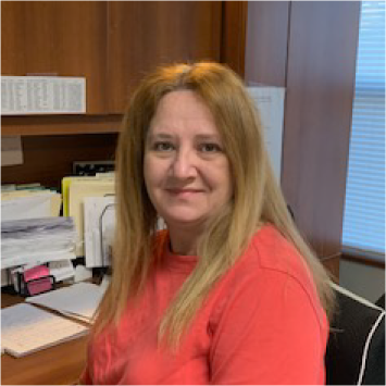 Woman with long blonde hair, wearing a red top, sitting at a desk in an office.