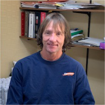 Man in blue shirt smiles, seated in front of shelves with binders and papers.