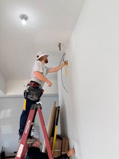 Two electricians working, one on a ladder installing wiring on a white wall.