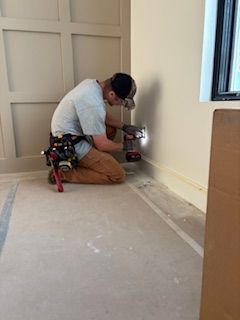 Carpenter installing trim, kneeling by a wall. Wearing a tool belt and cap, using a power drill on a cream-colored wall.