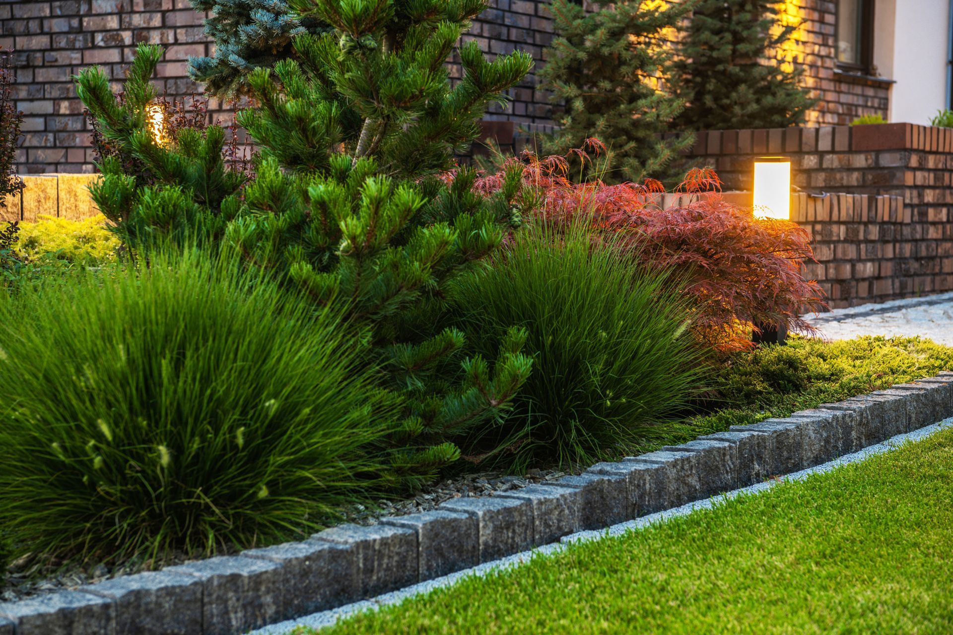 Landscaped front yard with green shrubs, brick border, and warm pathway lights beside a house
