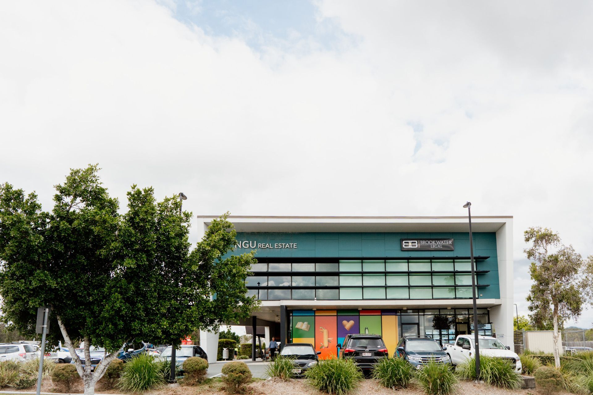 Modern glass-fronted building with green signage, trees, and shrubs in front
