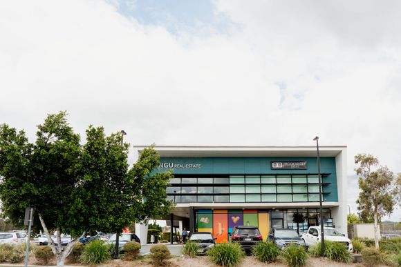Modern glass-fronted building with green signage, trees, and shrubs in front