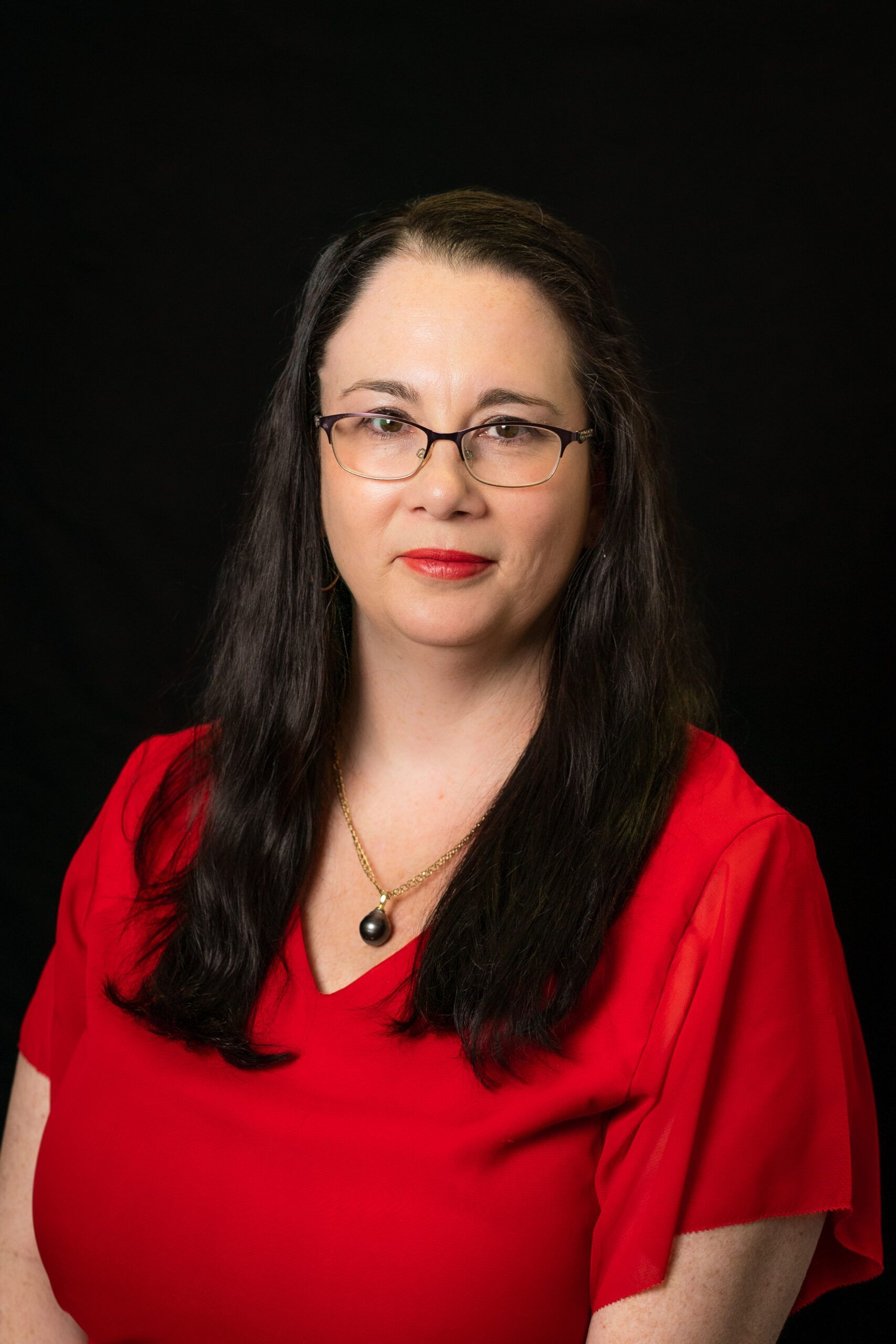 Woman with long dark hair and glasses wearing a red top against a black background