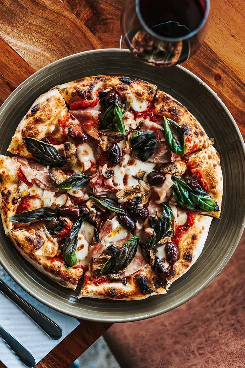 A Pizza is Sitting on a Plate on a Wooden Table Next to a Glass of Wine — Fedeles East Gosford in East Gosford, NSW