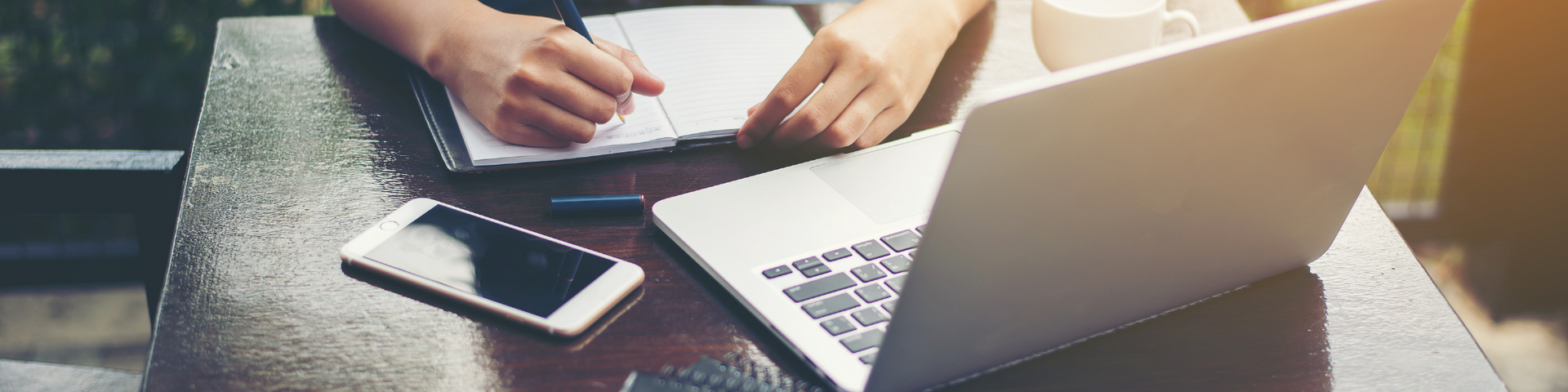 Hands typing beside a laptop, smartphone, and notebook on a desk in warm light