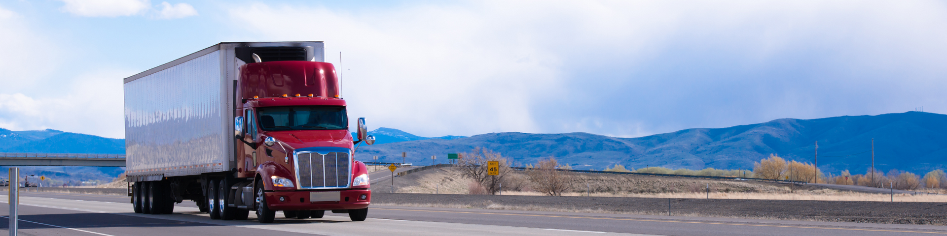 Red semi-truck pulling a white trailer on a desert highway with mountains in the background