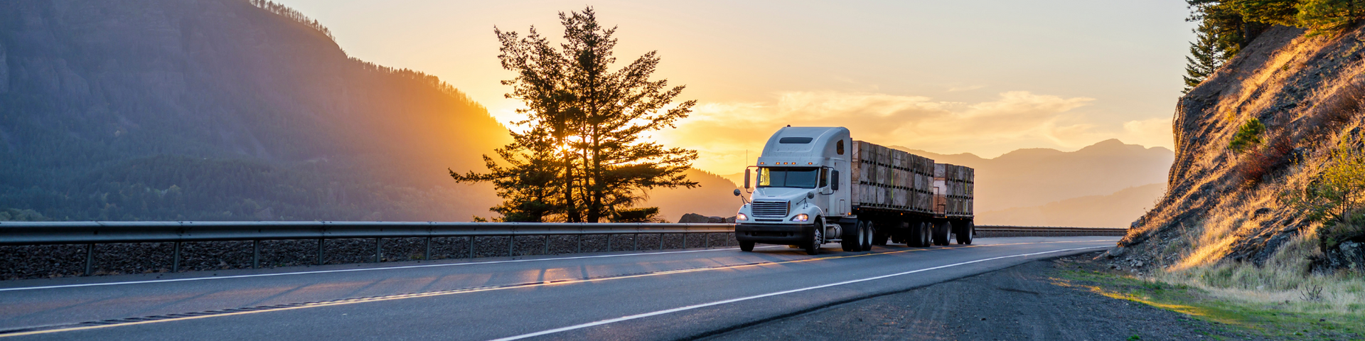 RV driving on a mountain road at sunset, with dramatic cliffs and a lone tree silhouetted by orange sky