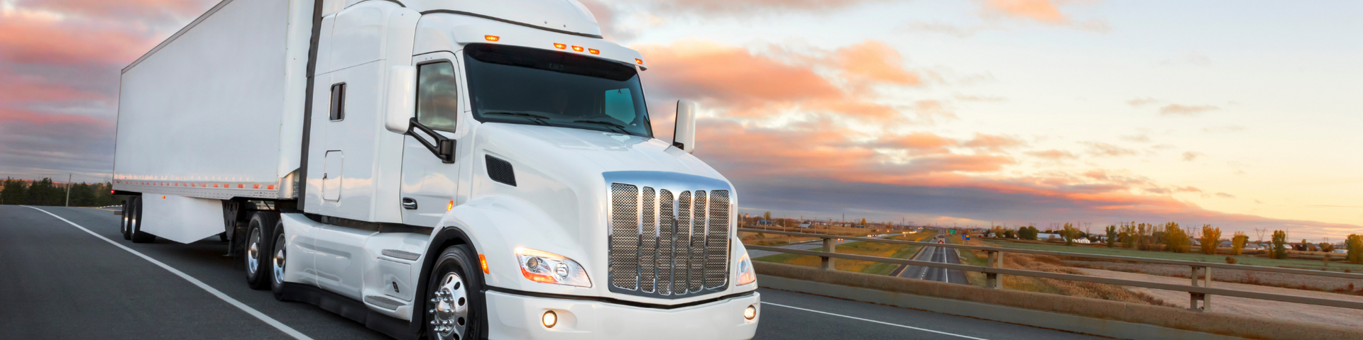 White semi-truck driving on a highway at sunset with a city skyline in the distance