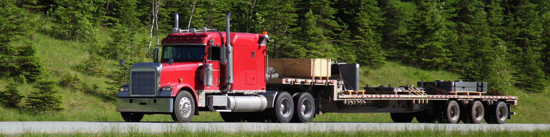 Red semi-truck hauling a flatbed trailer loaded with stacked lumber on a road by green trees