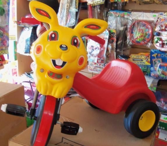 Triciclo amarillo con cara de conejo, asiento rojo y ruedas negras en el escaparate de una juguetería.