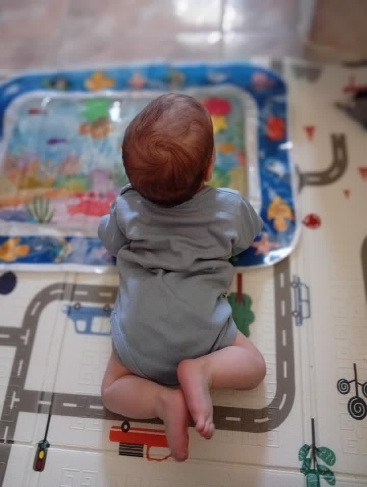 Un bebé sentado sobre una alfombra de juegos frente a una colorida bandeja de actividades llena de juguetes.