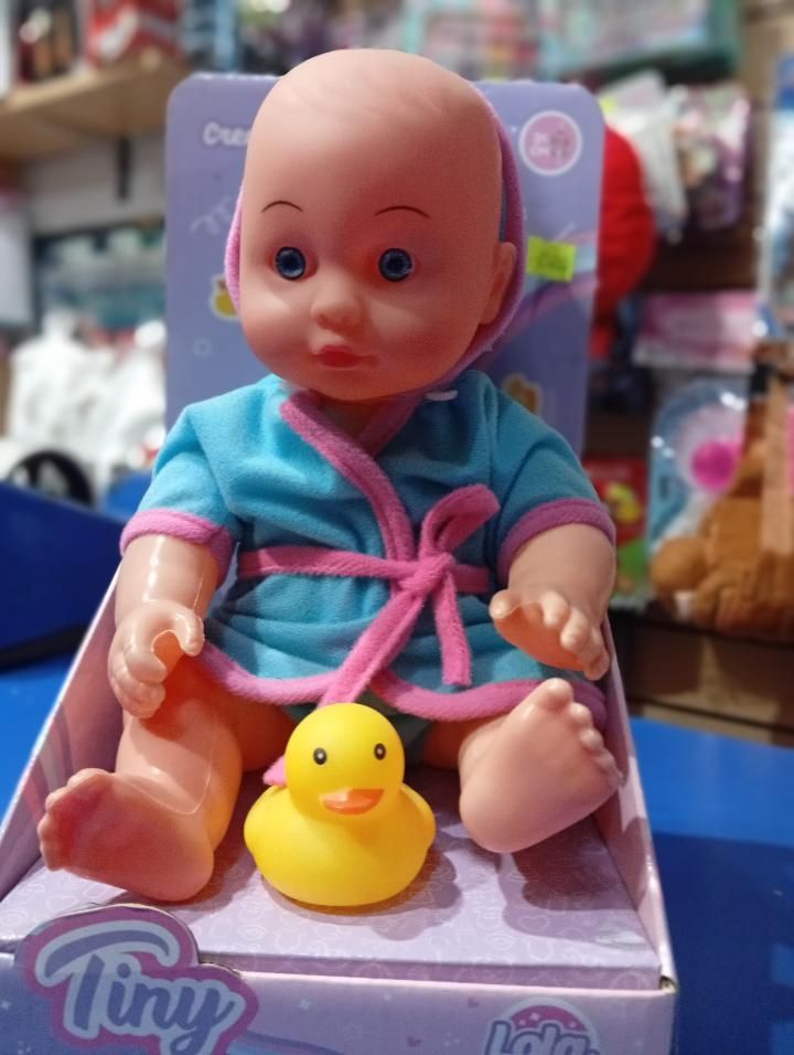 Muñeca bebé de juguete con una bata azul con ribete rosa, sentada en una caja junto a un pequeño patito de goma amarillo.