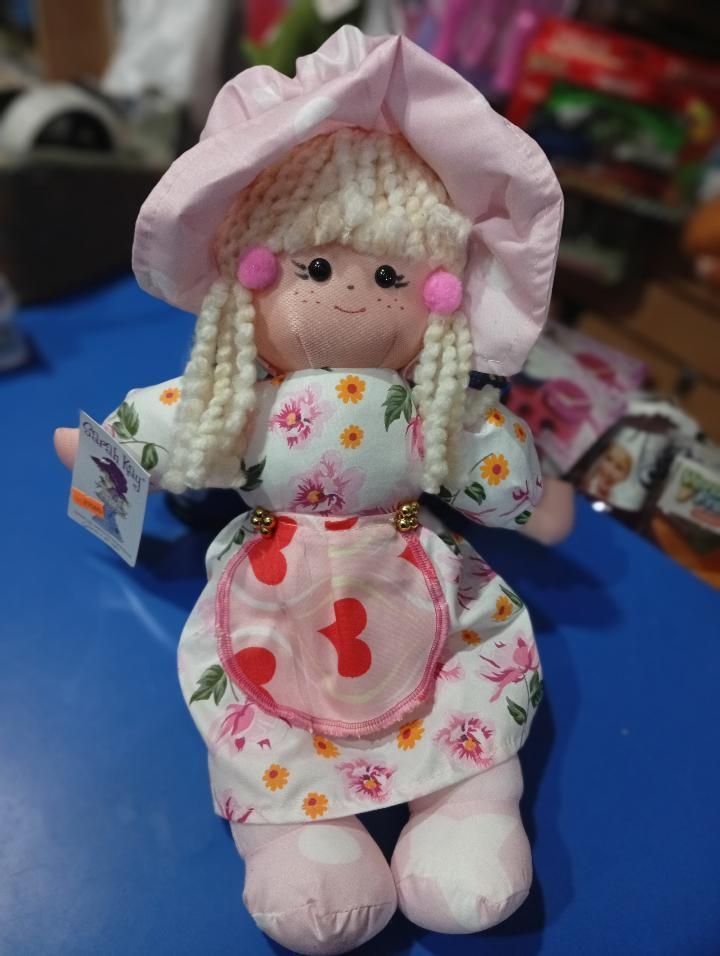 Muñeca de peluche con sombrero rosa y vestido floral, sentada sobre un fondo azul.