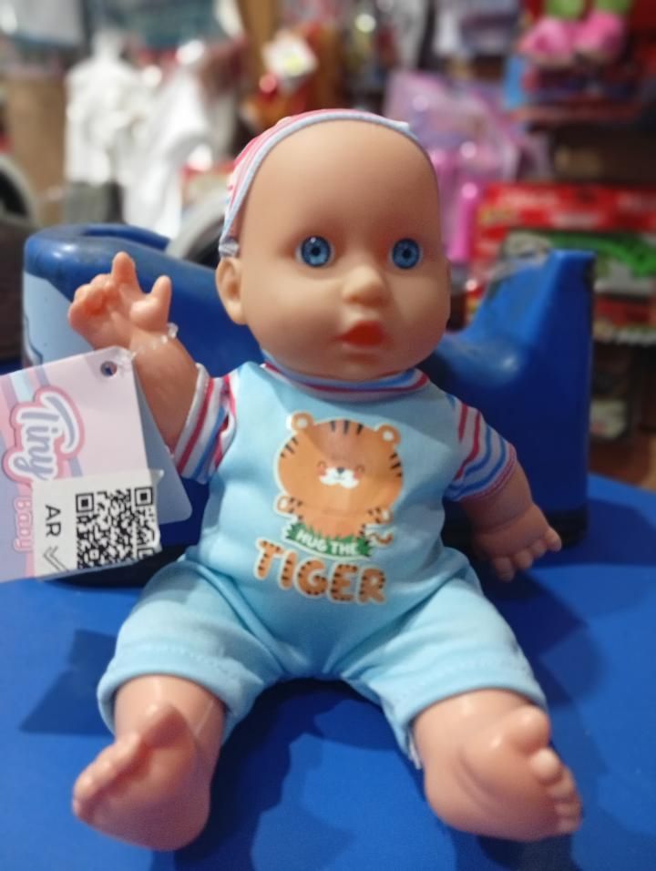 Muñeca bebé azul con un pelele de tigre azul claro sentada en una silla azul, sosteniendo una etiqueta con un código QR.