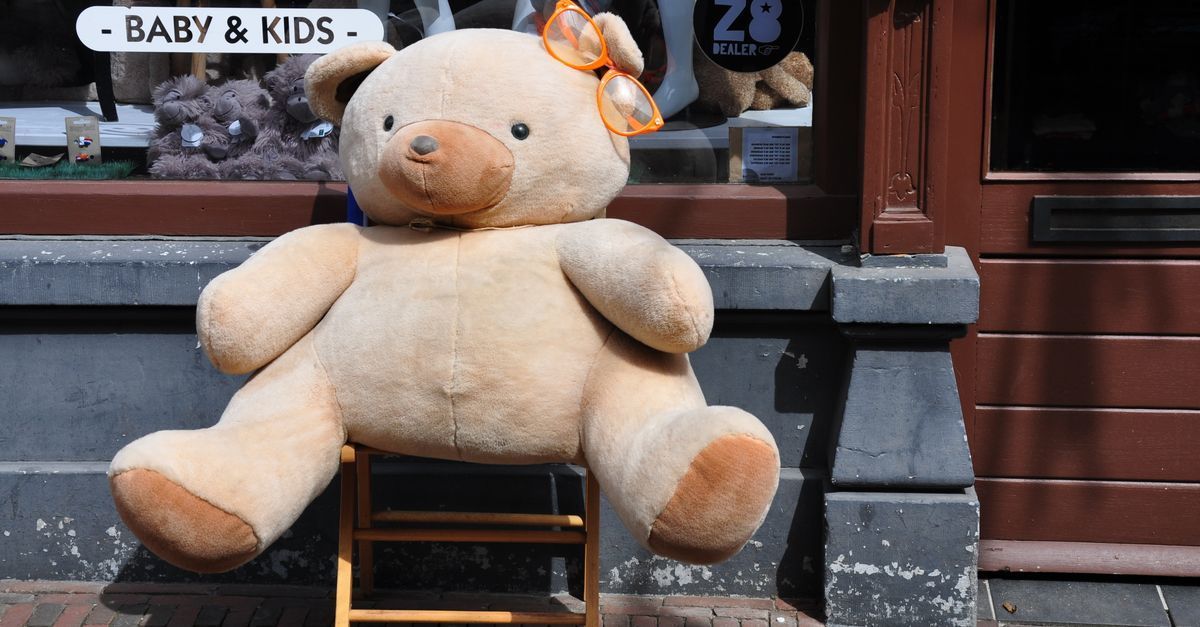 Un gran oso de peluche beige está sentado en un taburete frente al escaparate de una tienda con la etiqueta