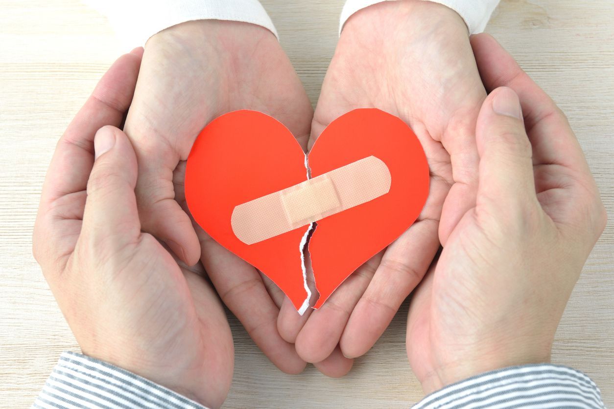 Two hands hold a red paper heart that is split in two and mended with an adhesive bandage.