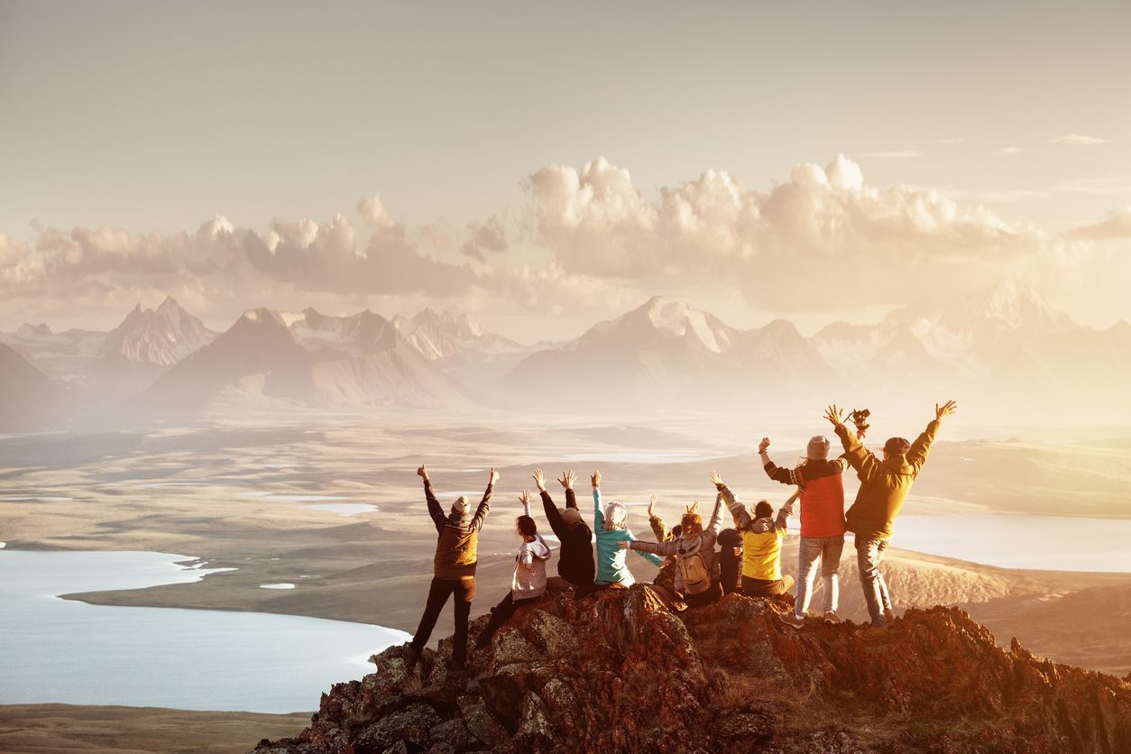 A group of hikers stands on a mountain peak at sunset, arms raised in celebration overlooking a lake and mountain range.