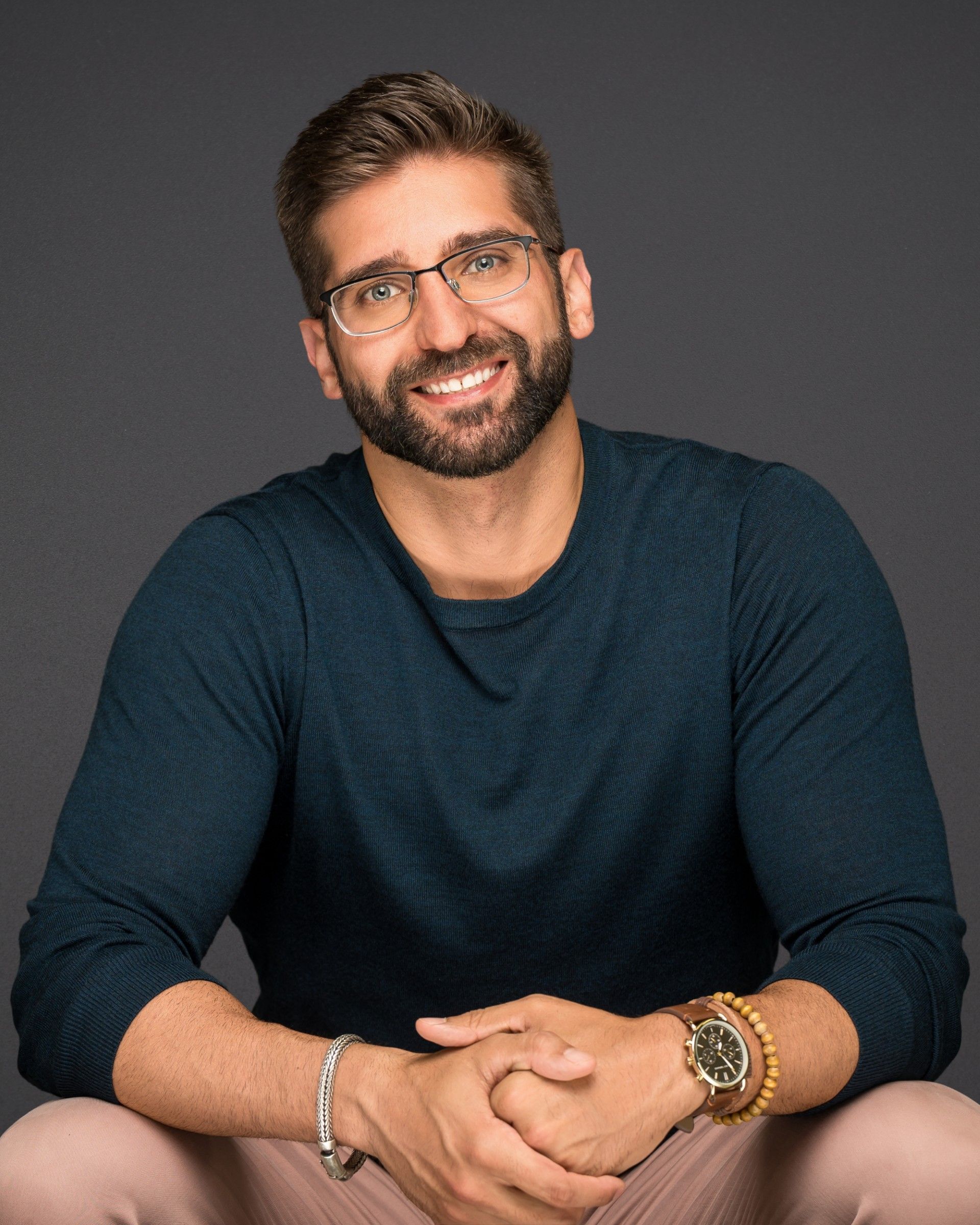 A smiling person with a beard and glasses wears a blue sweater against a dark gray background, hands clasped together.