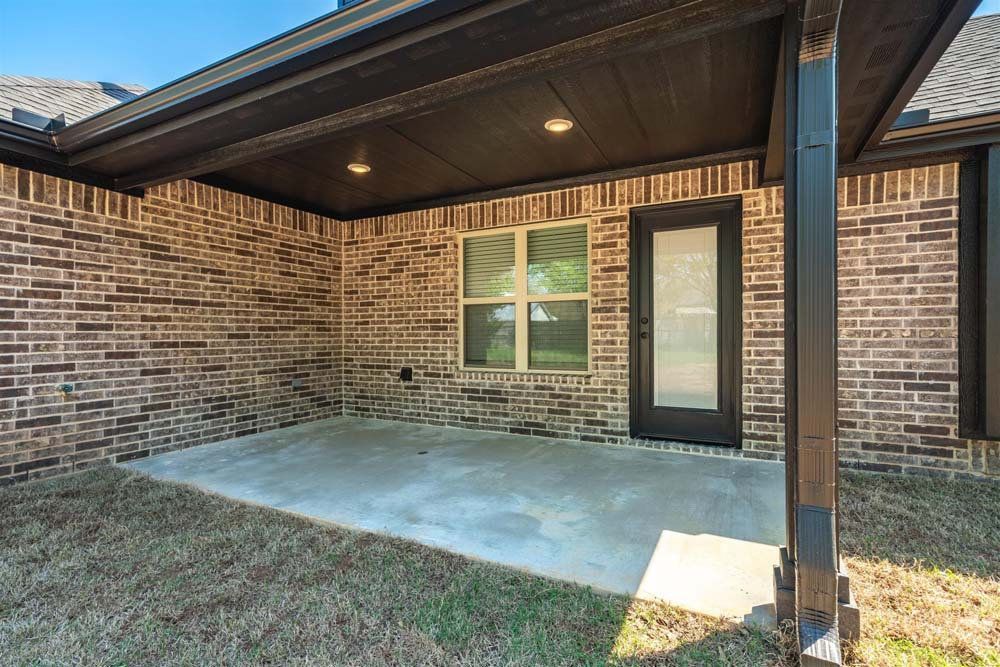 Covered brick patio with concrete slab, black support posts, and a glass back door under a roof overhang