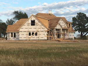Partially built suburban house with exposed framing and siding in a grassy field under a cloudy sky