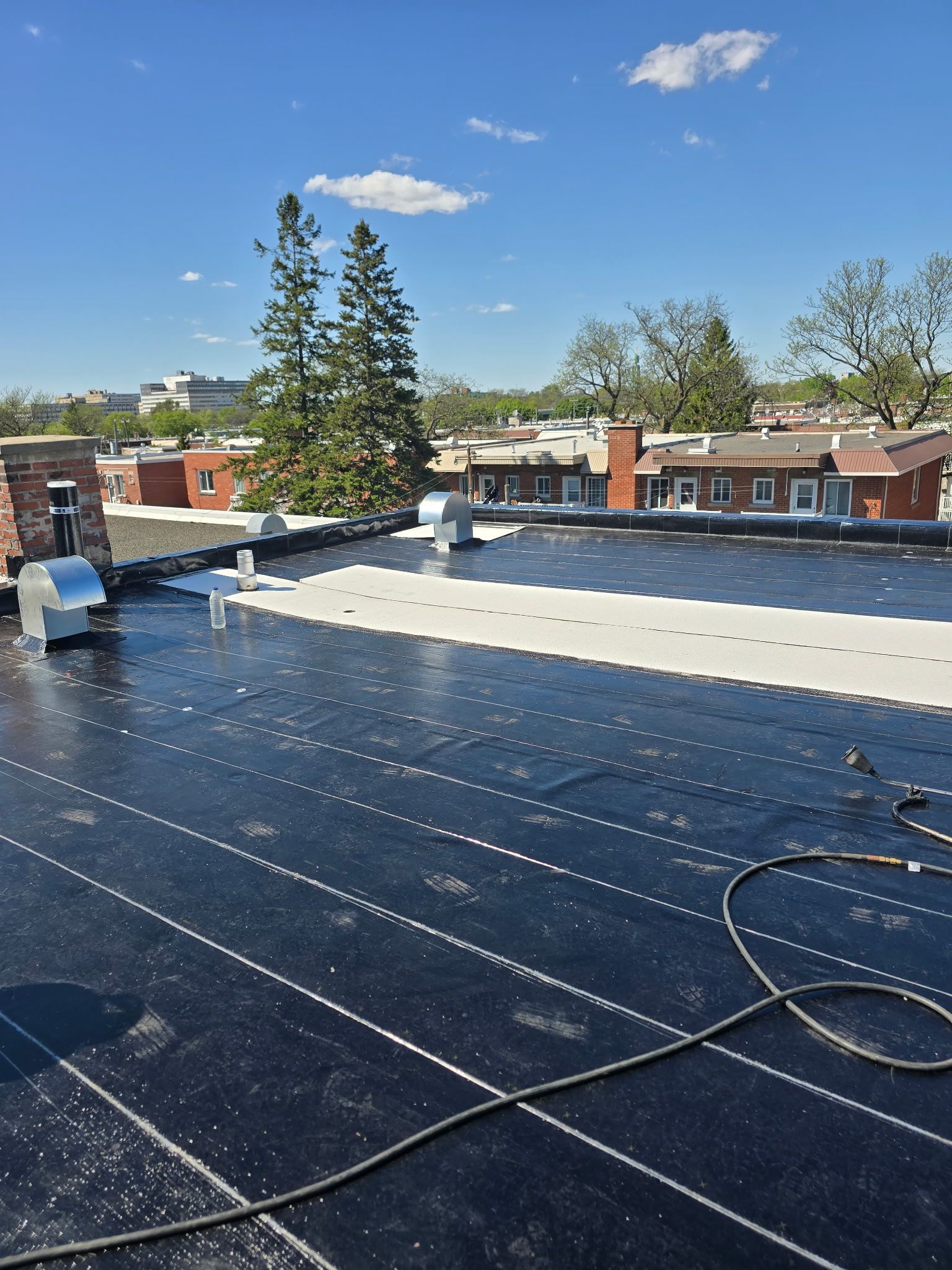 Toit plat sous un ciel bleu, avec une membrane d'étanchéité sombre, des tuyaux de ventilation et des maisons et des arbres à proximité.