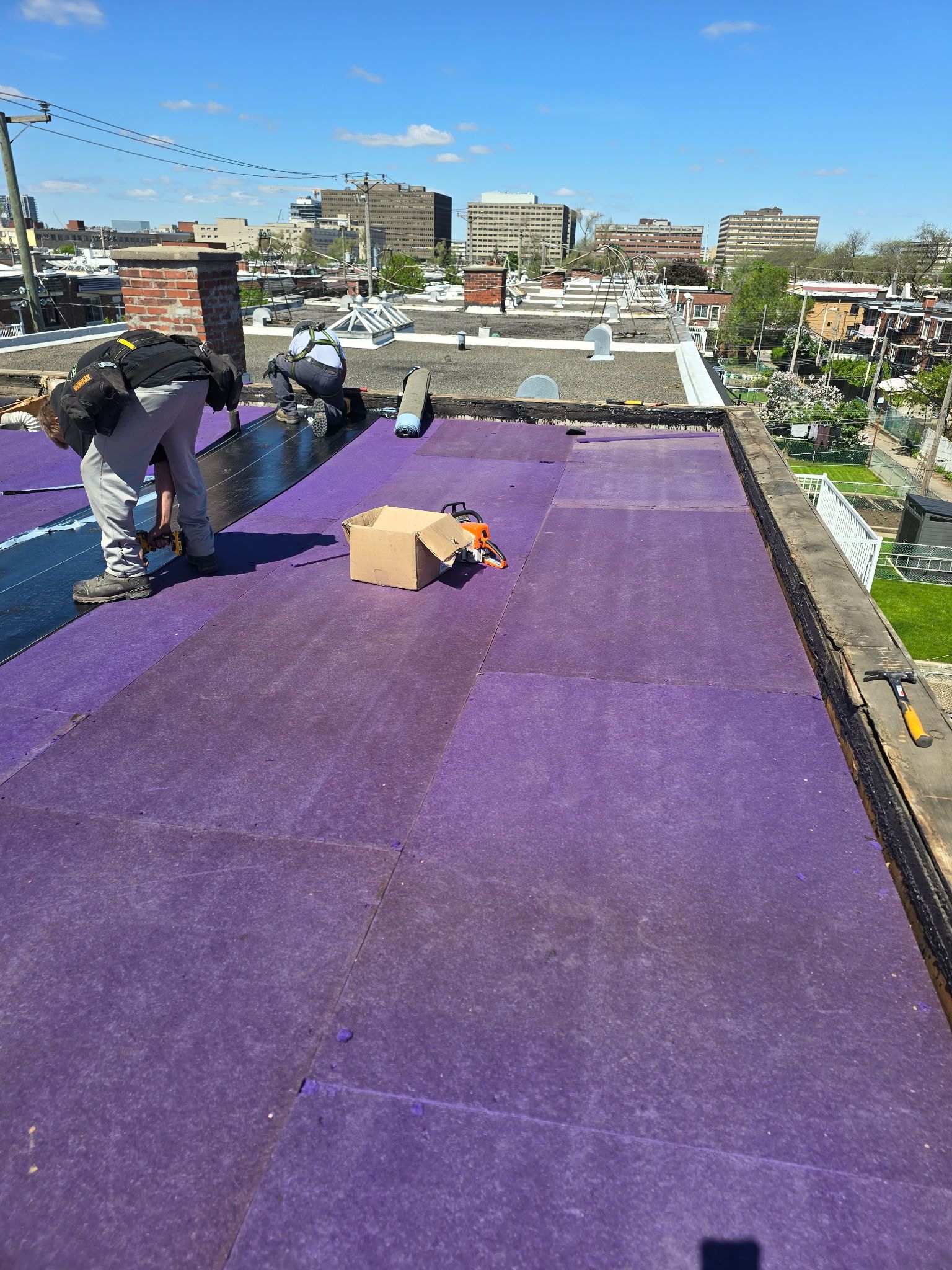 Des ouvriers installent une membrane d'étanchéité violette sur un toit, à côté d'une boîte en carton.