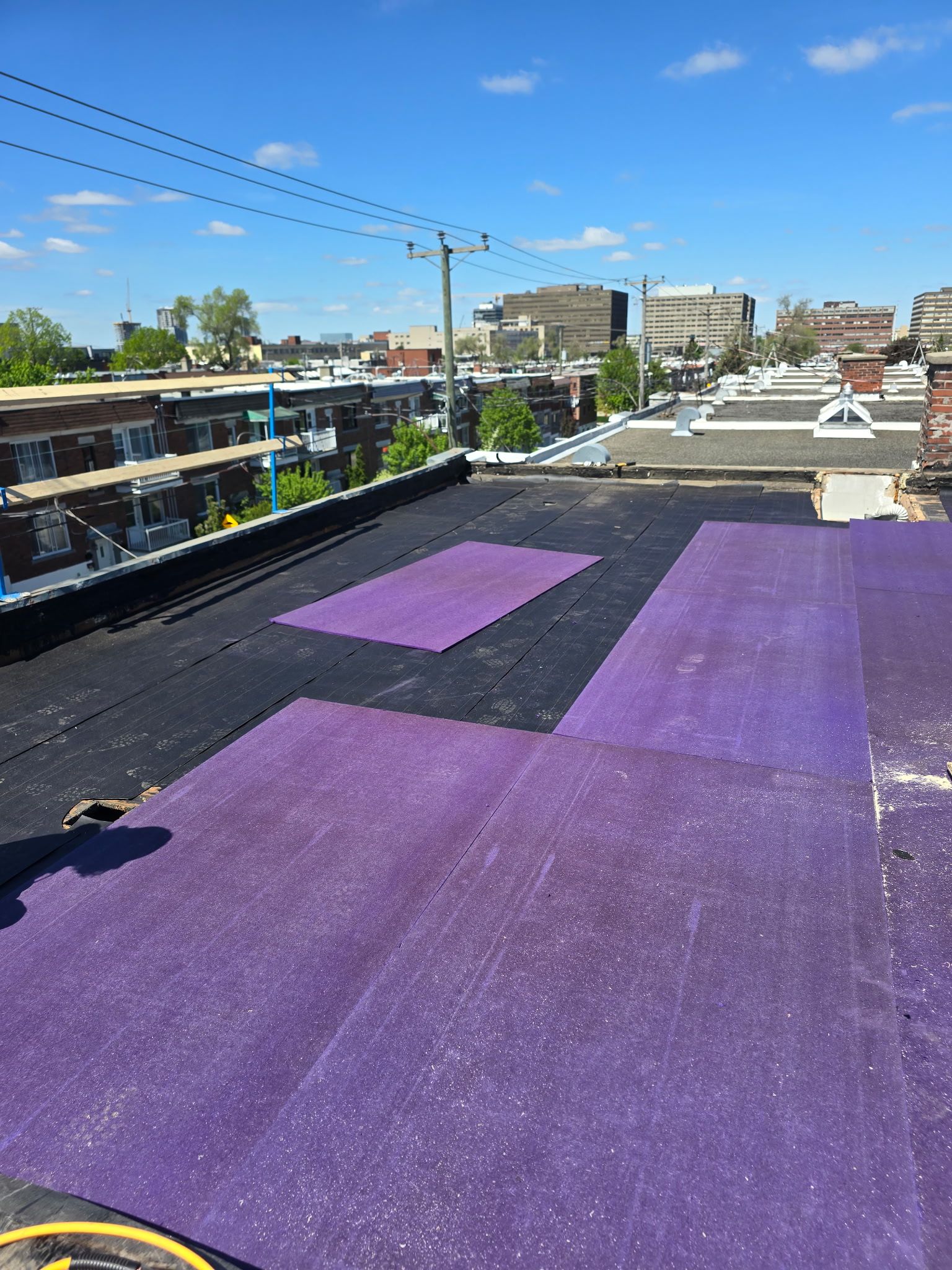 Toiture recouverte d'un matériau de couverture violet sous un ciel bleu ensoleillé, donnant sur les bâtiments et les lignes électriques environnantes.