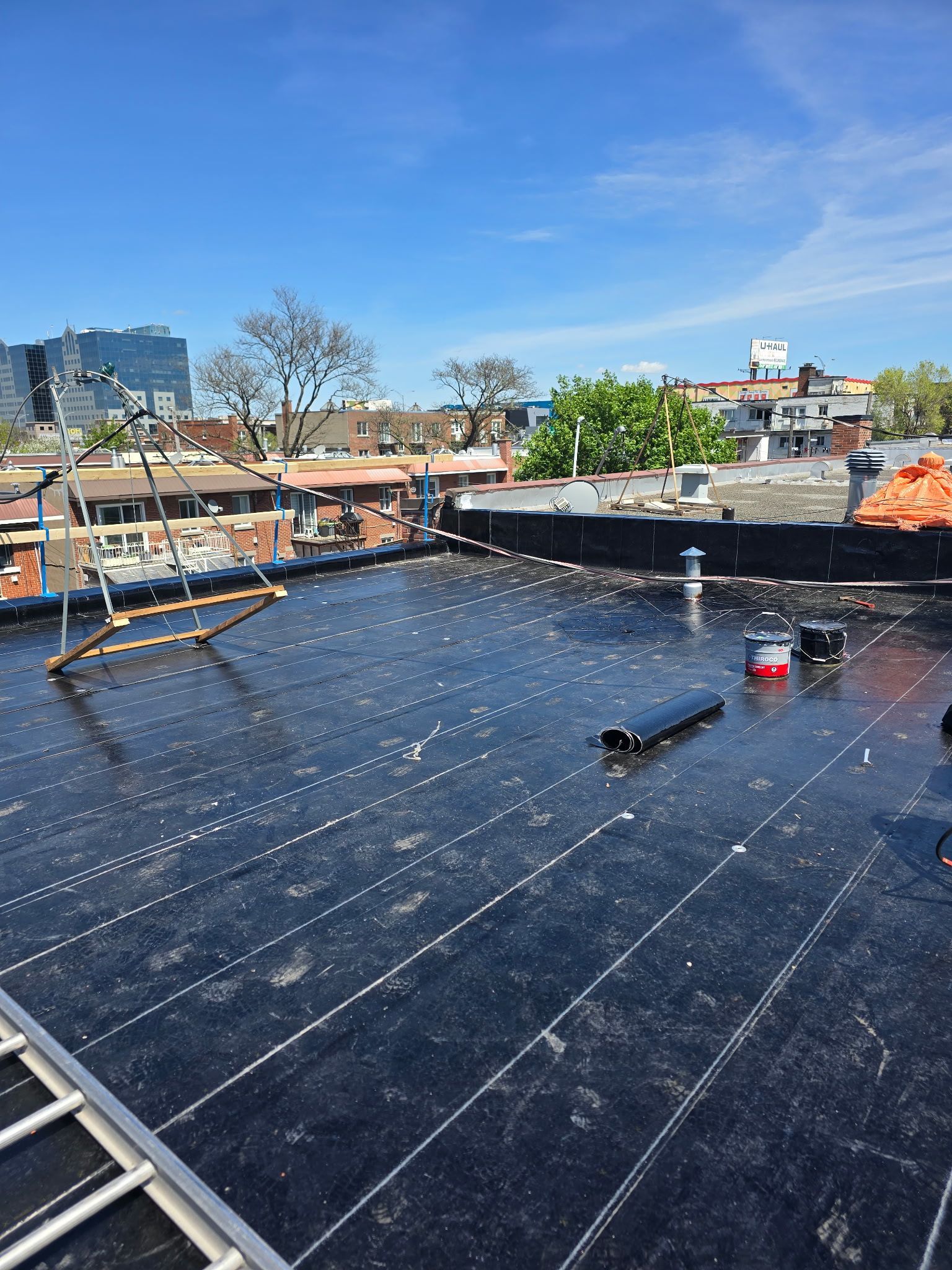 Toit recouvert d'une membrane d'étanchéité noire, de conduits de ventilation et de bâtiments urbains sous un ciel bleu.