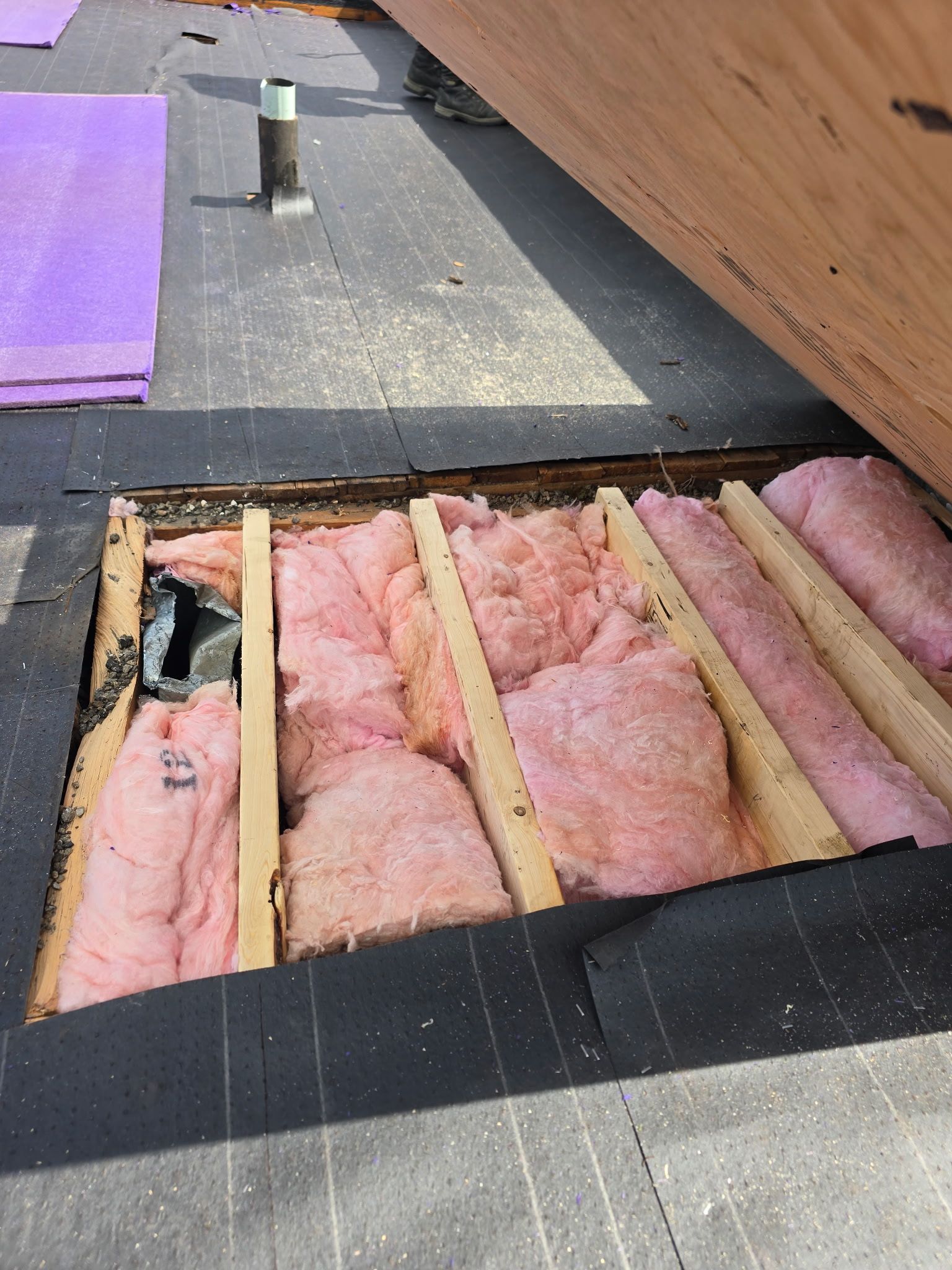 Toiture avec isolation rose apparente entre les solives en bois et un tuyau de ventilation sur feutre bitumé noir