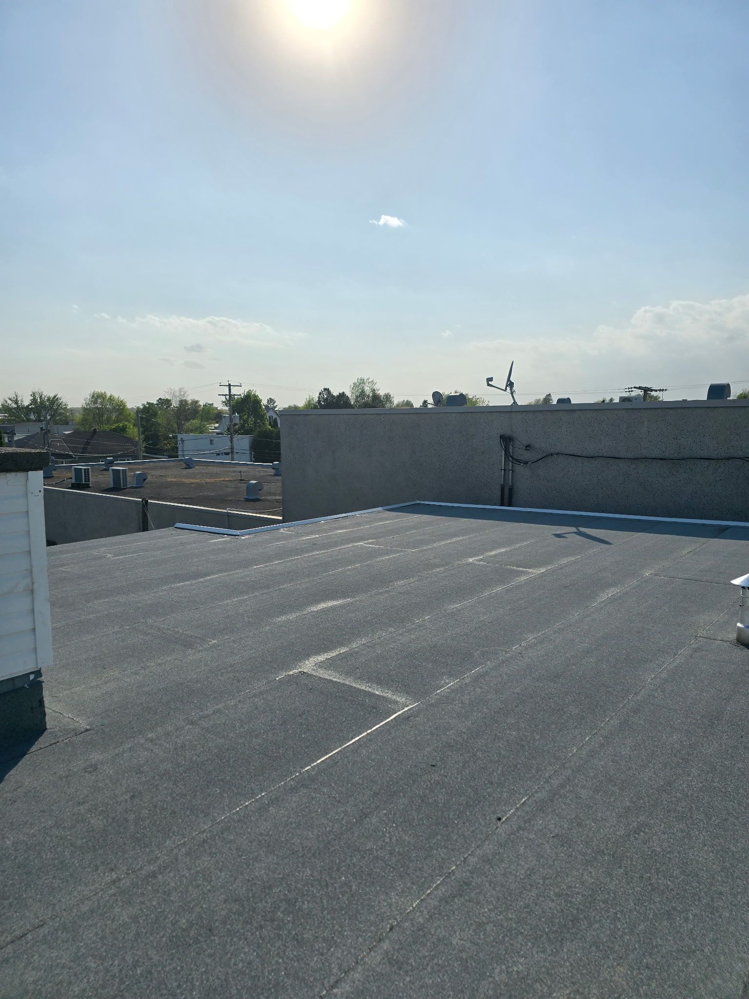 Toit-terrasse ensoleillé avec surface gravillonnée, murets blancs et arbres au loin sous un ciel bleu limpide.