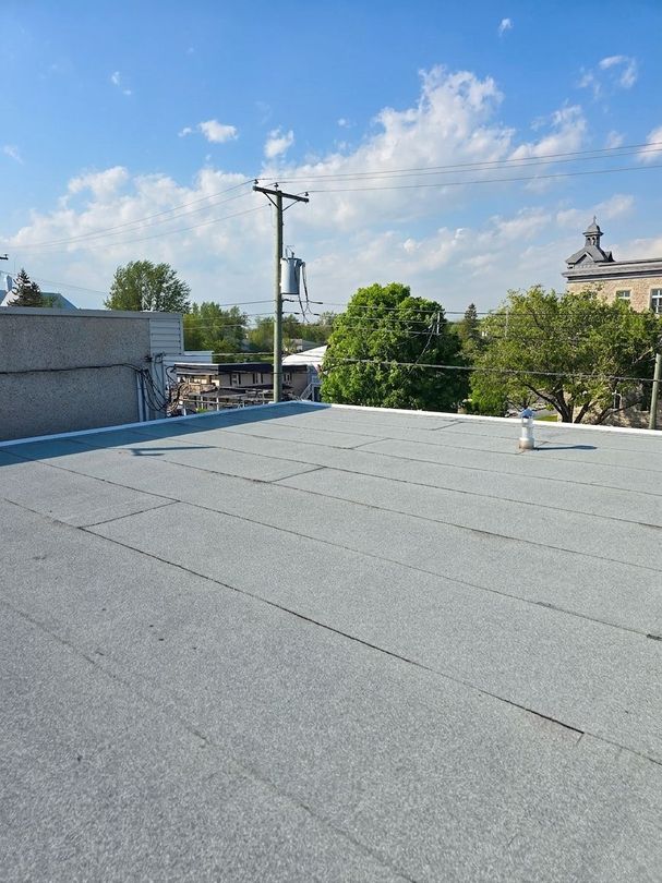 Toit plat gris sous un ciel bleu avec des nuages, des arbres, un poteau électrique et des bâtiments au loin