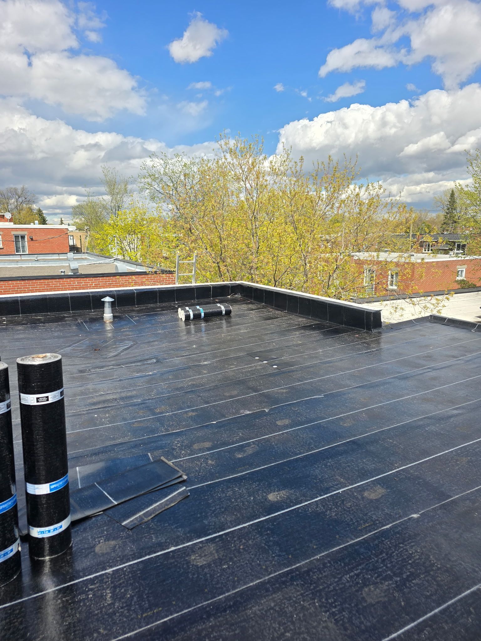 Toit-terrasse avec toiture plate noire, aérations et ciel bleu nuageux au-dessus des bâtiments voisins.