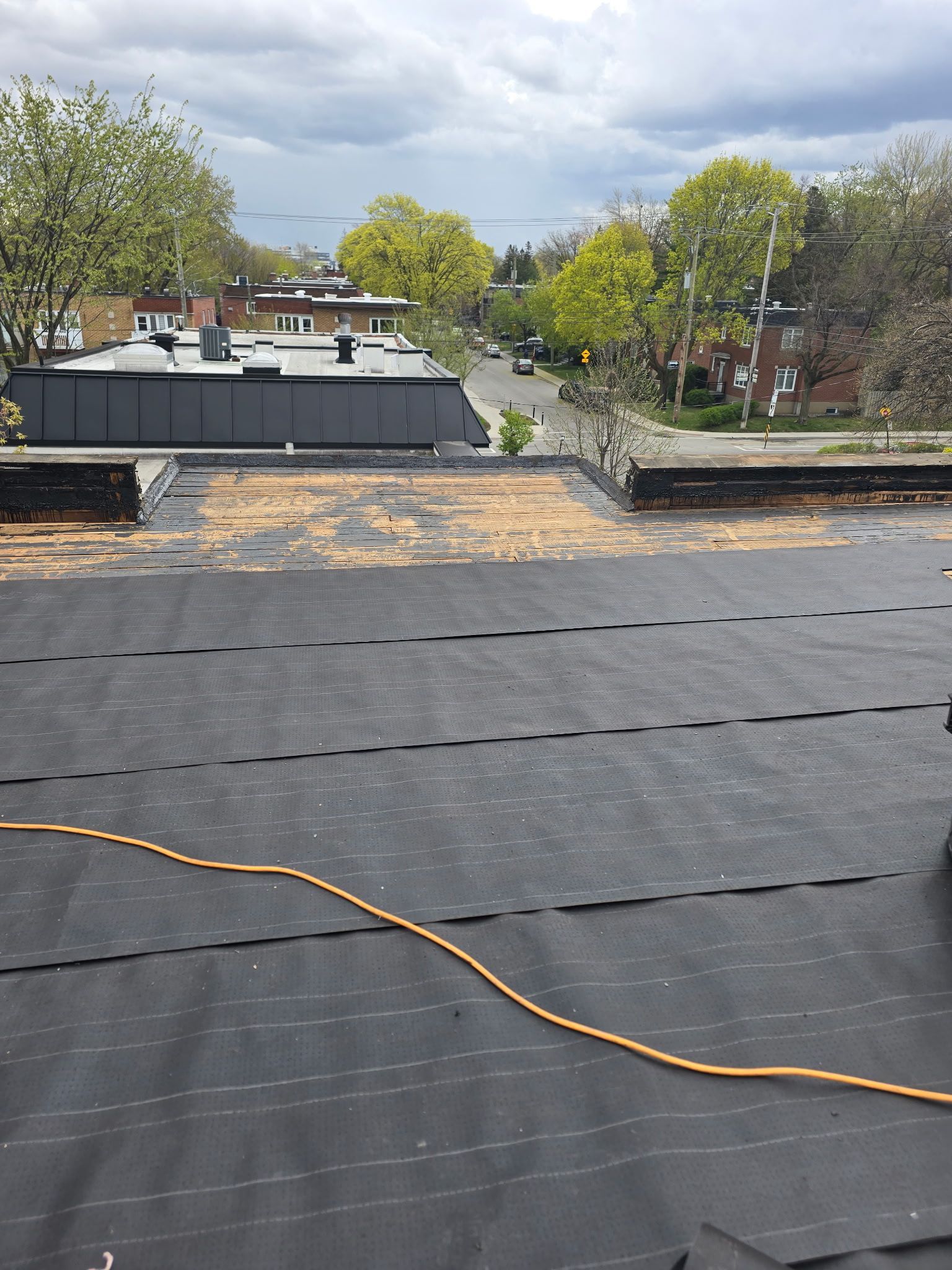 Toit noir plat en réparation, avec câble jaune, arbres et ciel nuageux en arrière-plan.